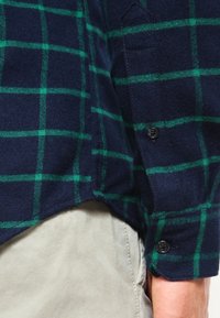 Camicia di flanella blu navy con motivo a quadri verdi, caratterizzata da polsini abbottonati e un orlo leggermente curvo. Texture morbida e vestibilità standard.