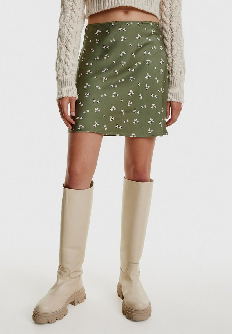 Groene bloemenrok met een gladde textuur, versierd met kleine witte bloemen. Gecombineerd met beige kniehoge laarzen met dikke zolen.