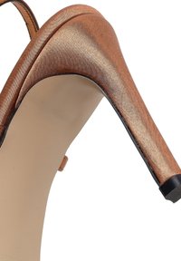 Primer plano de un zapato de tacón alto de color cobre brillante con tacón delgado y suela beige sobre un fondo blanco.