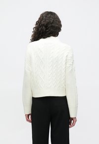 Suéter corto blanco de punto con cuello alto, que presenta un patrón texturizado y puños acanalados. Llevado sobre pantalones negros.