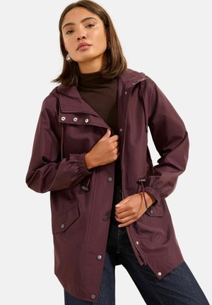 Frau trägt eine dunkelviolette Kapuzen-Regenjacke mit Druckknöpfen und Kordelzügen, kombiniert mit dunklen Jeans und einem schwarzen Rollkragenpullover.