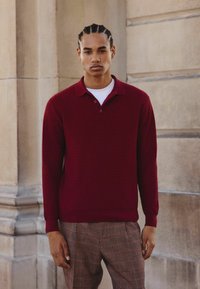 Pull en maille bourgogne avec un col et trois boutons, associé à un pantalon à carreaux marron, placé devant un fond en pierre.