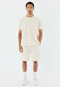 Camiseta de manga corta texturizada beige y pantalones cortos a juego. El modelo lleva zapatillas blancas y calcetines cortos con marca. Diseño simple y contemporáneo.