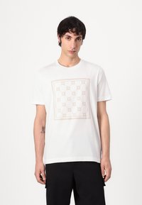 Camiseta blanca de algodón con un gráfico cuadrado que presenta un patrón floral repetido en tonos beiges. Mangas cortas y escote redondo.