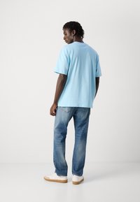 Άνδρας με σκούρο δέρμα και κοντά dreadlocks, φορώντας ένα γαλάζιο T-shirt, φθαρμένα γαλάζια τζιν και λευκά αθλητικά παπούτσια, μπροστά σε μια άδεια φόντο.