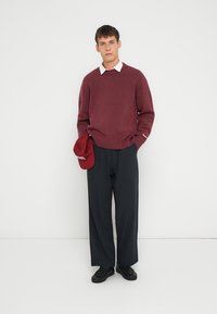 Suéter burdeos con dobladillo acanalado, camisa blanca con cuello debajo, pantalones de rayas navy, sosteniendo una gorra roja, zapatos negros. Fondo minimalista.