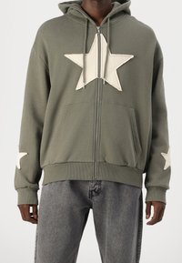 Felpa con zip verde oliva in cotone, caratterizzata da grandi applicazioni a stella color crema sul davanti e sulle maniche, con tasca a marsupio e polsini a costine.