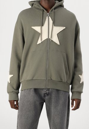 Sudadera con cremallera de color verde oliva, confeccionada en algodón, con grandes aplicaciones de estrellas color crema en el frontal y las mangas, con un bolsillo tipo canguro y puños acanalados.