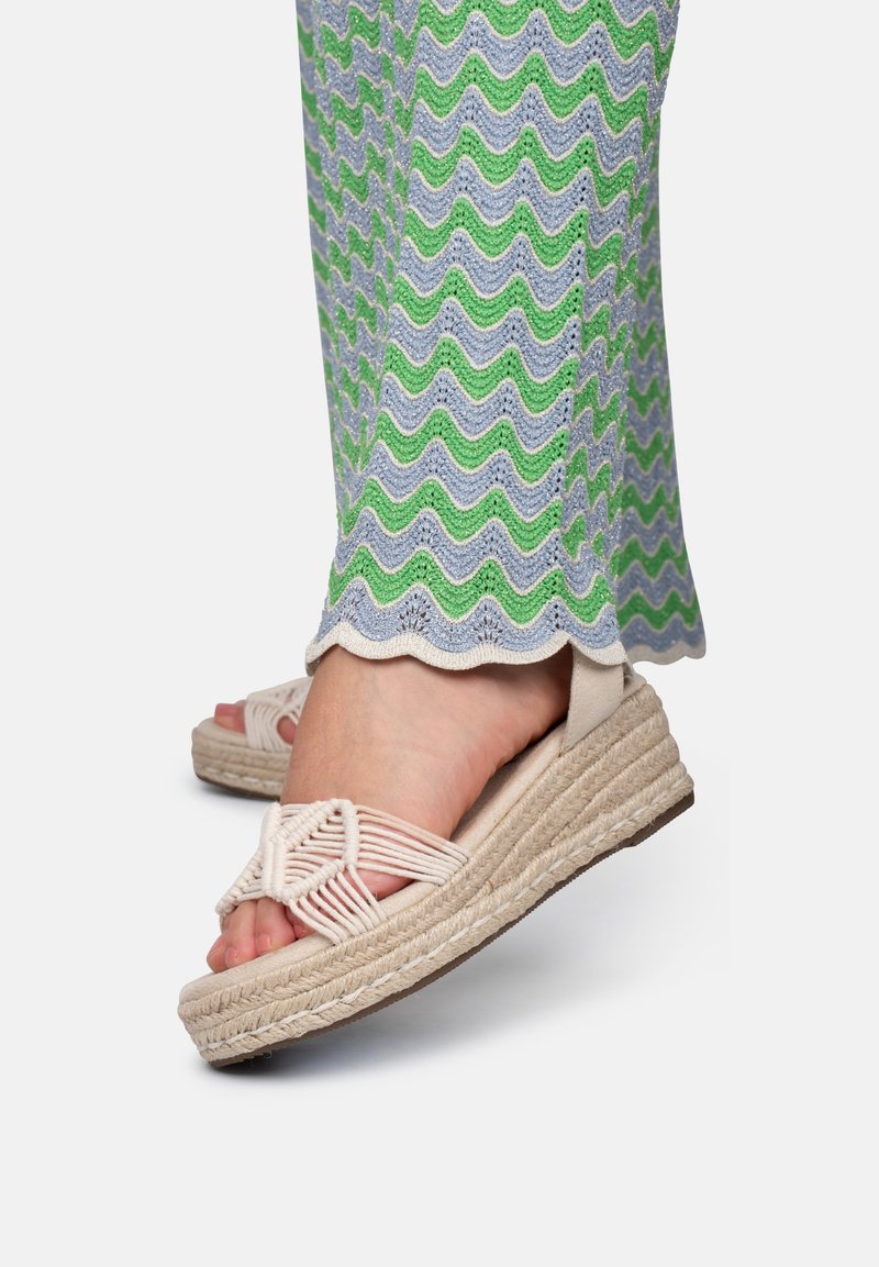 Espadrille-sandalen met een geweven lichtbeige bovenkant, een jute-wrapped wigzool en een textuurpatroon op zachte pastelgroene en grijze broeken.