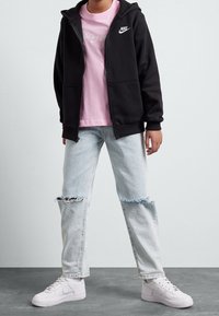 Persona vistiendo una sudadera con cremallera negra de Nike, una camiseta rosa de Nike, jeans claros desgastados y zapatillas blancas de Nike, de pie sobre un suelo gris.