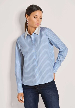 Frau trägt ein blau-weiß gestreiftes Hemd mit weißem Kragen und dunkle Jeans, steht mit einer Hand auf der Hüfte.