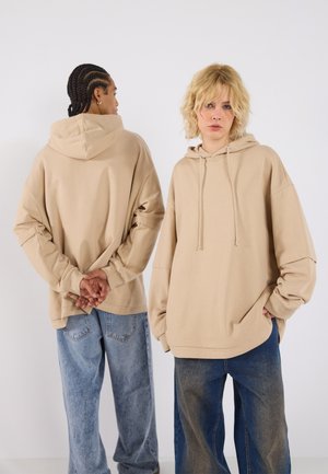 Oversized beige hoodie with a drawstring hood, front kangaroo pocket, and relaxed sleeves, paired with light blue oversized jeans.