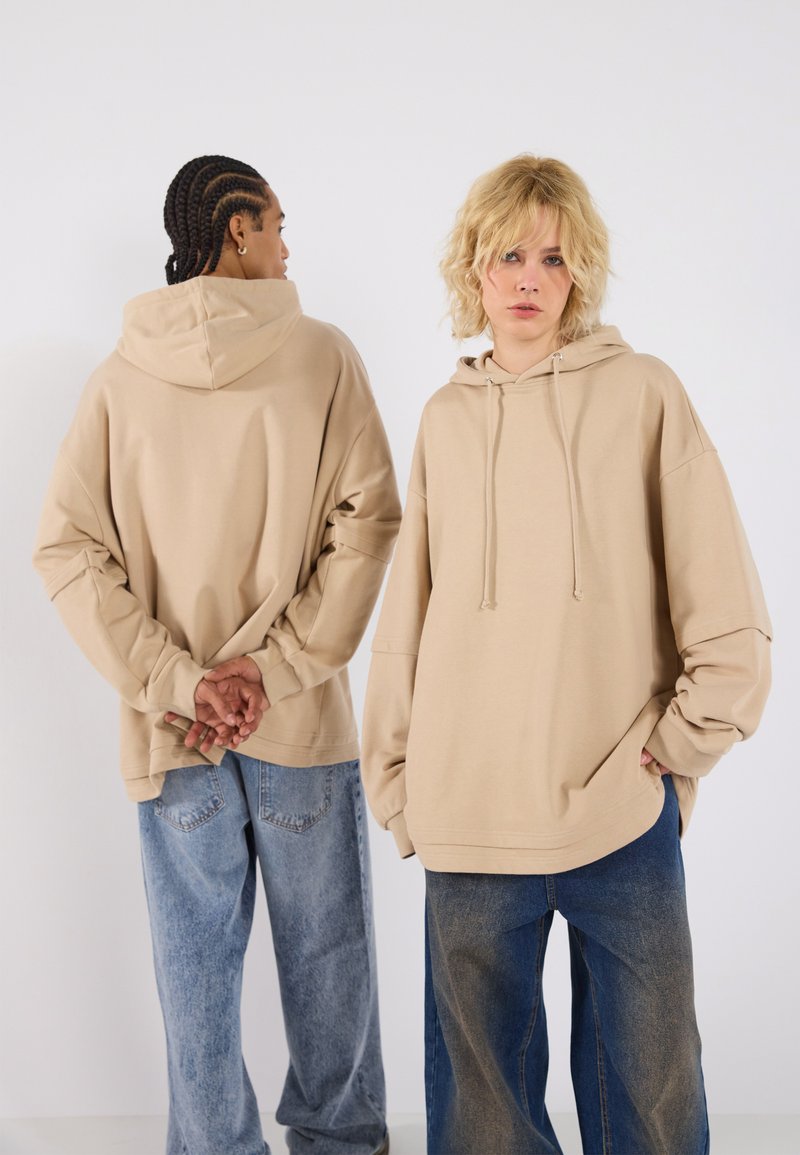 Oversized beige hoodie with a drawstring hood, front kangaroo pocket, and relaxed sleeves, paired with light blue oversized jeans.