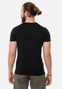 Schwarzes kurzärmliges T-Shirt, enganliegendes Design, weicher Stoff, Rundhalsausschnitt, sichtbare Naht, kombiniert mit hellgrünen Hosen. Rückansicht.