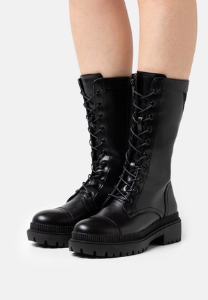 Bottes de combat en cuir noir à design montantes jusqu'au genou, avec lacets sur le devant et semelles robustes, présentant une texture lisse et une quincaillerie solide.