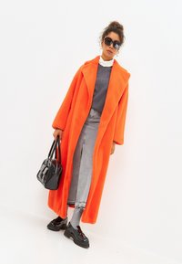 Palton lung din blană artificială portocalie, cu reveruri late, purtat peste un pulover gri și o fustă din denim. O geantă neagră și pantofi negri lucioși completează look-ul.