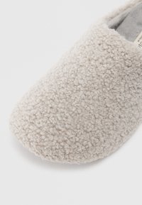 Beige pantoffel gemaakt van zacht, textuur materiaal. Ronde neus en slip-on ontwerp met een pluche interieur voor comfort.