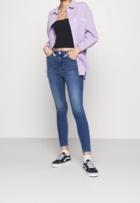 Person in einem helllila Button-Up-Hemd über einem schwarzen Croptop, mit blauen Skinny-Jeans, weißen Socken und schwarz-weißen Turnschuhen, die Hände in den Taschen.