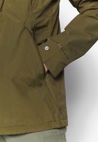 Olivgrön jacka med en slät yta och dragkedjefickor; har "Omni-Tech"-logotyp på ärmen och en elegant, modern design.