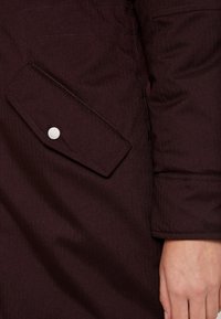 Primo piano di una giacca color bordeaux con tessuto strutturato, tasca laterale con bottone a pressione argento e dettagli cuciti per maggiore durata.