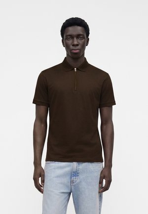 Hombre con cabello corto y rizado, vestido con una camiseta polo de manga corta marrón oscuro y vaqueros azul claro, de pie frente a un fondo blanco liso.