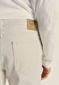 Jean en denim clair avec une poche arrière visible et une étiquette rectangulaire marron, affichant des symboles d'entretien et le nom de la marque.
