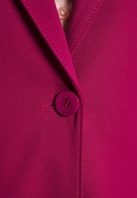 Gros plan d'un blazer magenta avec un seul bouton assorti fermé près du revers et des détails de couture visibles.
