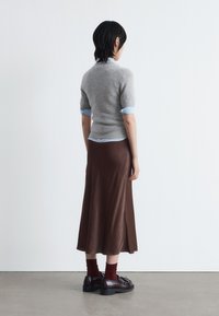 Grauer Kurzarm-Pullover über einem hellblauen Hemd mit Kragen, kombiniert mit einem braunen Satinrock und schwarzen Schuhen. Rote Socken sind sichtbar.