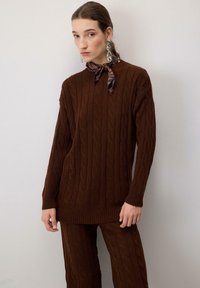 Ruskea kaapelikudottu neulepaita pitkillä hihoilla, jossa on ribbireunat hihoissa ja helmassa. Yhdistetty yhteensopiviin ruskeisiin leveälahkeisiin housuihin.