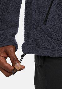 Giacca in fleece blu scuro con superficie texturizzata, dotata di un regolatore con cordino all'orlo. Include una tasca con zip sul petto.