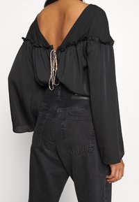 Mujer vistiendo una blusa negra de manga larga con la espalda descubierta y detalle de lazo, combinada con jeans de mezclilla negros de cintura alta, vista desde atrás.