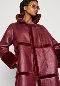 Bordeaux faux læderjakke med høj krave, lynlås lukning og blødt teksturerede detaljer på ærmer og krop, der har en boxy form og strukturerede paneler.
