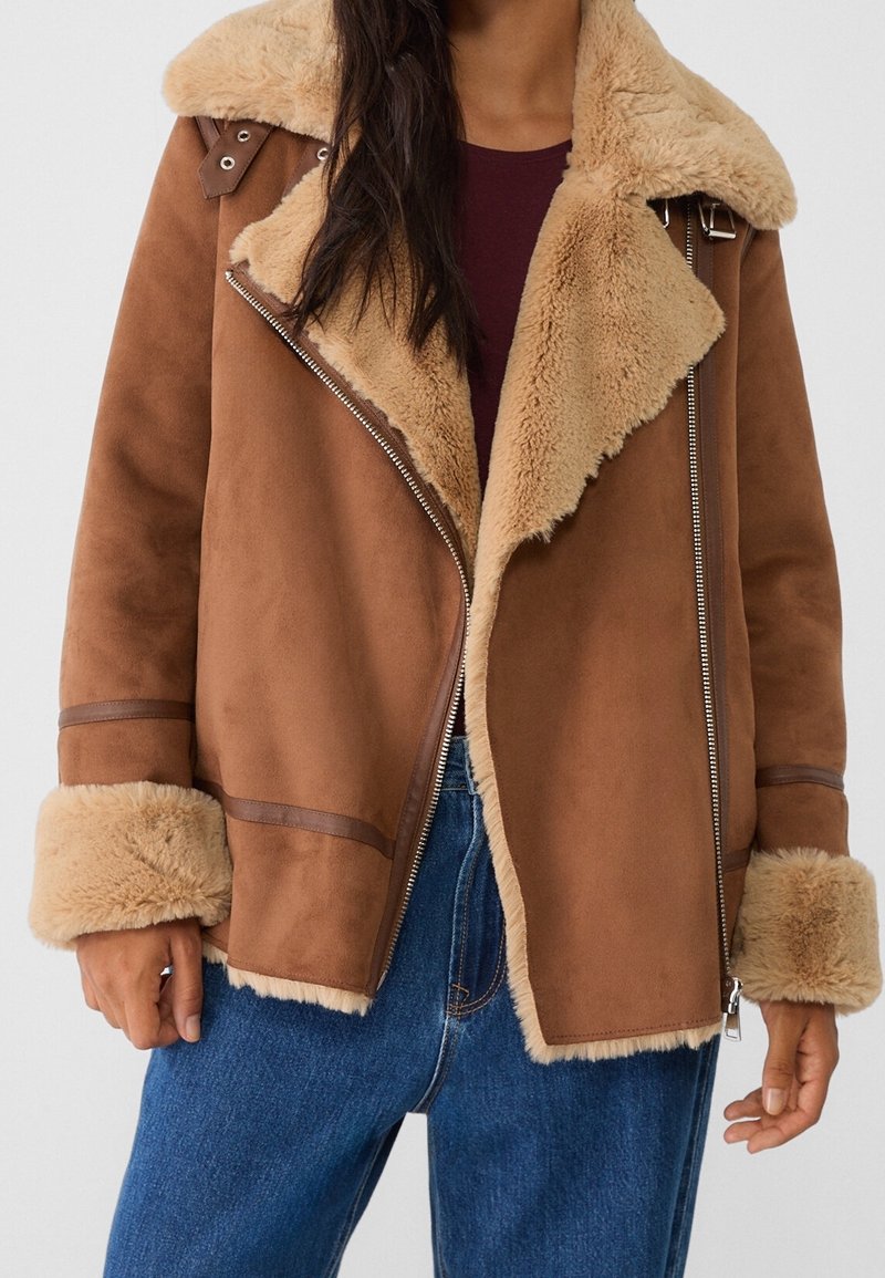 Femme portant une veste en shearling marron avec un grand col, des détails à fermeture éclair et un jean bleu sur un fond uni.