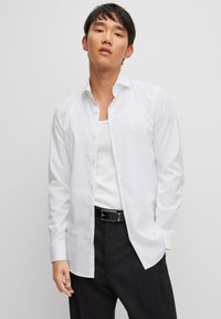Camisa blanca de manga larga con frente abotonado, cuello clásico y textura suave, combinada con una camiseta blanca básica y pantalones negros.