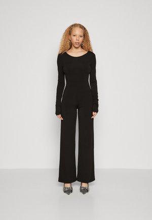 Combinaison noire à manches longues, corps ajusté, pantalons à jambes larges et ouvertures pour les pouces. Associée à des chaussures texturées noires et argentées.