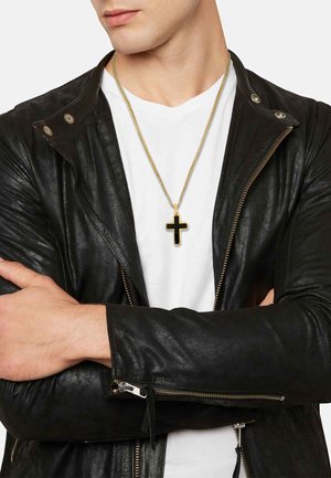 Gouden ketting met een zwart kruis hanger, gedragen over een wit T-shirt en onder een zwarte leren jas met ritsmouwen.