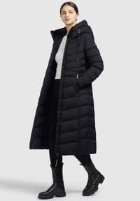 Cămașă puffer lungă, neagră, cu glugă, având un design matlasat, fermoar în față și buzunare laterale. Purtați peste un top alb și pantaloni negri.