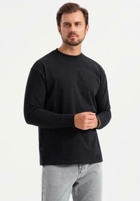 Mann mit Bart, der ein schwarzes langärmliges Shirt und hellgraue Jeans trägt, steht mit gefalteten Händen vor einem schlichten weißen Hintergrund.