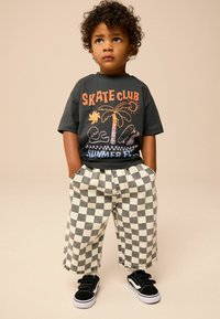Gray t-shirt with "SKATE CLUB" graphic, palm tree, and waves; oversized beige and gray checkered shorts; black sneakers with Velcro straps.