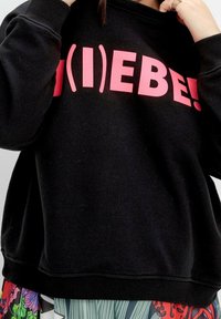 Czarny sweter z różowym napisem graficznym "(I)EBE!" z przodu. Posiada ściągacze przy rękawach i luźny krój, zestawiony z wzorzystymi spodniami.