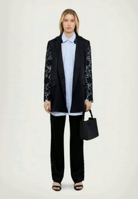 Czarna marynarka z koronkowymi rękawami, jasnoniebieska koszula, czarne spodnie i mała czarna torebka. Czarny sandały na odkrytej stopie. Gładkie tekstury, dopasowany krój.