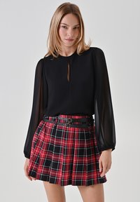 Schwarze Bluse mit langen Ärmeln und Ausschnitt in Form eines Schlüssellochs, kombiniert mit einem rot-schwarzen karierten, plissierten Minirock. Schwarzer Gürtel unterhalb der Taille.
