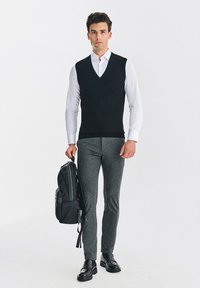 Czarny bezrękawnik na białej koszuli, w zestawie z szarymi dopasowanymi spodniami, czarnym plecakiem i czarnymi skórzanymi butami. Prosty design.