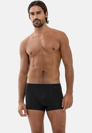 Muskelöser Mann mit dunklen Haaren und Bart steht und trägt schwarze Boxershorts vor schlichtem weißem Hintergrund.