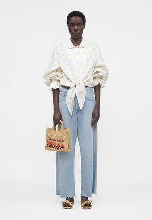 Γυναίκα που φοράει λευκό πουκάμισο με κίτρινα σχέδια δεμένο στη μέση, ανοιχτό μπλε παντελόνι με φαρδιά πόδια, καφέ σανδάλια, κρατώντας μια μικρή πλεκτή τσάντα με την επιγραφή «Vanessa Bruno».