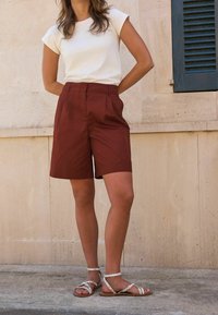 Shorts marron ajustés avec des plis associés à un t-shirt beige clair et des sandales blanches à lanières, le tout sur un fond neutre.