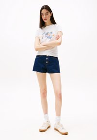 Witte graphic t-shirt met bloemmotief, gecombineerd met blauwe denim shorts met een knopenfront. Model draagt witte sneakers en sokken.
