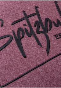 Schwarze gummierte Buchstaben "Spitzbul" auf einem strukturierten kastanienbraunen Hintergrund, mit einem fetten Schriftdesign, das ausgeprägte Kurven und Akzente aufweist.