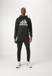 Czarna bluza Adidas z dużym białym logo, w połączeniu z czarnymi dresami i białymi butami sportowymi. Trzyma hantle w jednej ręce.