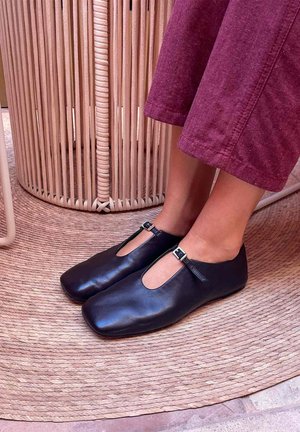 Pieds chaussés de chaussures noires en cuir à bride en T avec bouts carrés, portées avec un pantalon violet raccourci, debout sur un tapis rond tressé près d'une table d'appoint en osier.